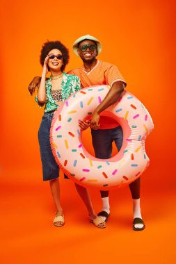 Full length of smiling young african american man in sunglasses and panama hat hugging stylish best friend and holding swim ring on orange background, friends in trendy casual attire clipart
