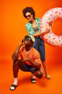 Full length of young african american woman in sunglasses and stylish outfit holding swim ring while standing near best friend in panama hat on orange background, friends in trendy casual attire clipart