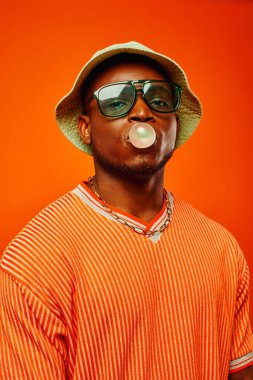 Portrait of trendy young african american man in sunglasses and panama hat blowing bubble gum and looking at camera while standing isolated on red, man with sense of style, confidence clipart