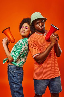 Confident young african american best friends in trendy summer outfits holding loudspeakers and looking at camera while standing isolated on red, friends in fashionable outfits clipart