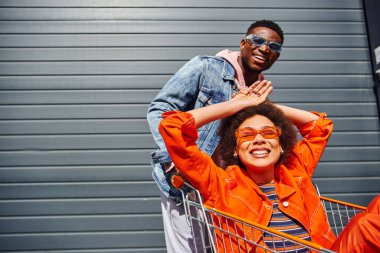 Cheerful young african american woman in sunglasses and bright outfit sitting in shopping cart near stylish best friend and building on urban street, friends hanging out together, friendship clipart