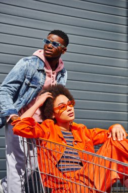 Fashionable young african american woman in sunglasses and bright outfit sitting in shopping cart near confident best friend in denim jacket and building at background, friends hanging out together clipart