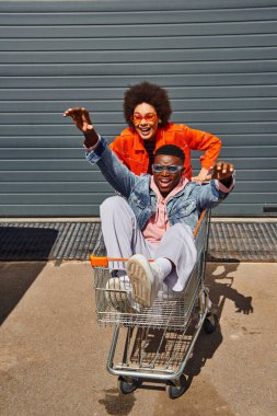 Cheerful african american woman in bright sunglasses and outfit having fun with best friend sitting in shopping cart near building on urban street, friends with stylish vibe concept clipart