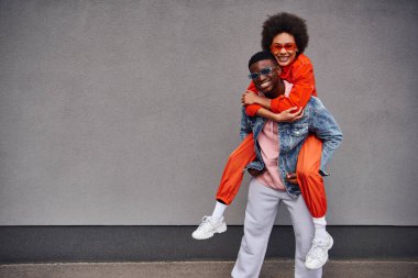 Smiling young african american woman in sunglasses and bright outfit piggybacking on stylish best friend and looking at camera near building on urban street, trendy friends in urban settings clipart