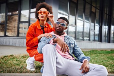 Stylish young african american woman in bright clothes wearing sunglasses and looking away near confident best friend sitting on border on urban street, stylish friends enjoying company clipart