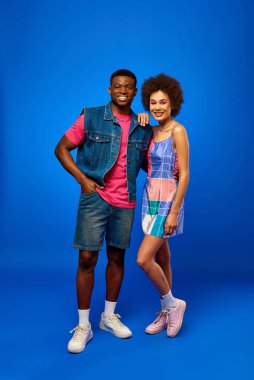 Full length of trendy and cheerful young african american best friends in summer clothes posing and looking at camera while standing on blue background, stylish friends radiating confidence clipart