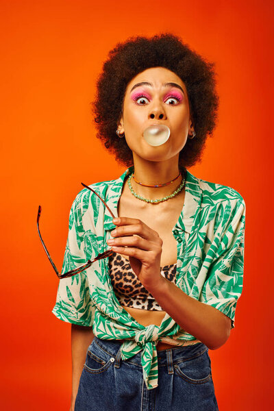 Portrait of excited young african american woman with bold makeup wearing summer outfit holding sunglasses and blowing bubble gum isolated on red, woman with sense of style, confidence