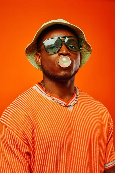 Portrait of trendy young african american man in sunglasses and panama hat blowing bubble gum and looking at camera while standing isolated on red, man with sense of style, confidence
