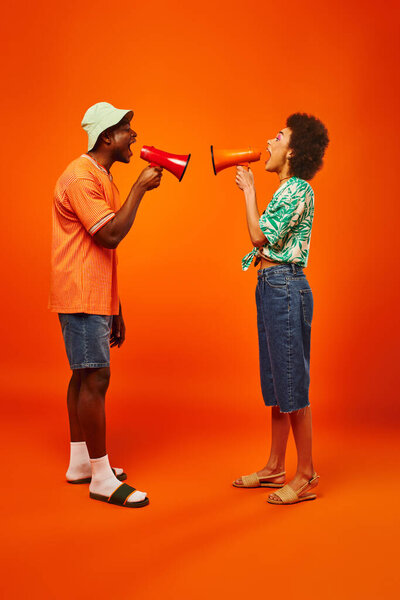 Side view of excited young african american best friends in stylish summer outfits screaming while holding loudspeakers while standing on red background, friends in fashionable outfits