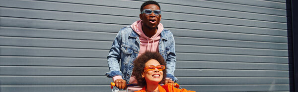 Excited young african american man in sunglasses and denim jacket standing near positive best friend sitting in shopping cart and building at background, friends hanging out together, banner 