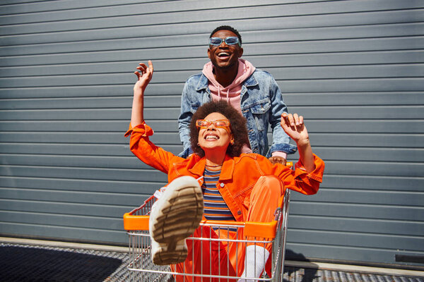 Cheerful young african american best friends in sunglasses and bright outfits having fun with shopping cart and spending time near building at background, friends hanging out together