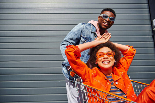 Cheerful young african american woman in sunglasses and bright outfit sitting in shopping cart near stylish best friend and building on urban street, friends hanging out together, friendship