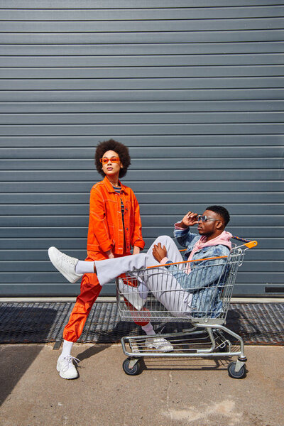 Full length of fashionable young african american man wearing sunglasses while sitting in shopping cart near best friend in bright clothes and building outdoors, friends with stylish vibe
