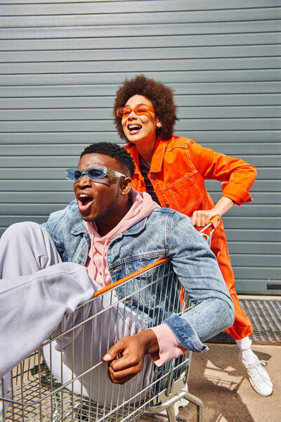 Smiling young african american woman in bright outfit and sunglasses having fun with stylish and scared best friend sitting in shopping cart near building on urban street, friends with stylish vibe