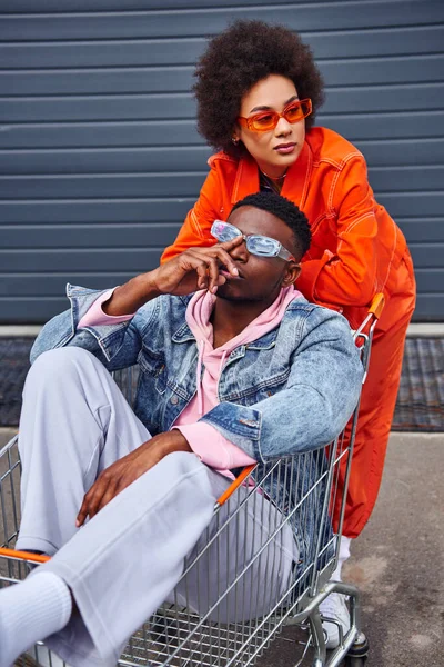 stock image Modern young african american woman in sunglasses and bright outfit looking away while standing near best friend sitting in shopping cart and building on urban street, stylish friends in city