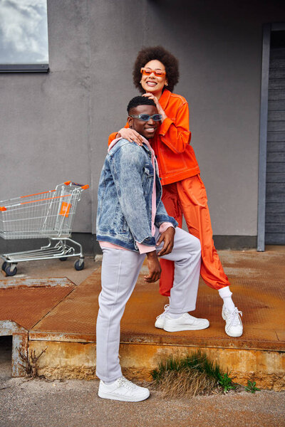 Full length of cheerful young african american woman in bright outfit and sunglasses hugging best friend in denim jacket while standing on stairs near building outdoors, stylish friends in city