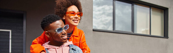 Positive young african american woman in sunglasses and modern outfit hugging best friend and looking away while standing together near building on urban street, stylish friends in city, banner 