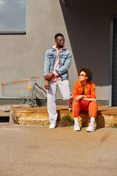 錆びた階段の近くに位置するサングラスや明るい衣装で流行の若いアフリカ系アメリカ人の親友の完全な長さ都市の通りの背景に建物 都市の概念のスタイリッシュな友人 — ストック写真