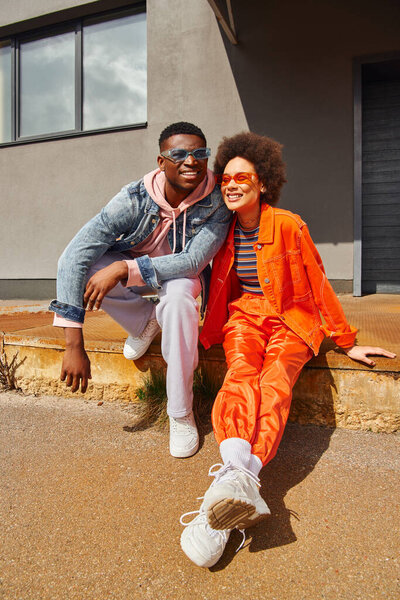 Smiling young african american best friends in sunglasses and bright clothes sitting on rusty stairs near building at background and spending time on urban street, stylish friends in city
