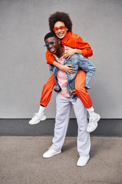 stock image Full length of cheerful and stylish young african american woman piggybacking on best friend in sunglasses and denim jacket while standing near building outdoors, trendy friends in urban settings