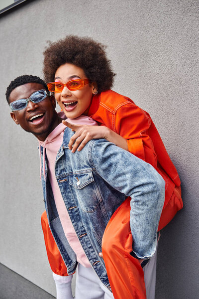 Cheerful young african american woman in stylish sunglasses pointing with finger while piggybacking on best friend and standing near building on urban street, friends with trendy aesthetic