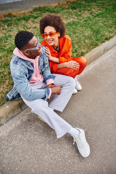 stock image High angle view of positive and modern african american woman in sunglasses and bright outfit talking to best friend while sitting together on border on urban street, friends with trendy aesthetic