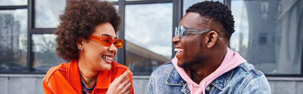 Smiling young african american woman in sunglasses and bright outfit talking to best friend in denim jacket while spending time on urban street, friends with trendy aesthetic, banner 
