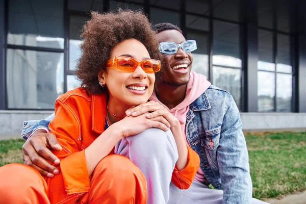 stock image Pleased young african american woman in sunglasses and bright outfit looking away while spending time with best friend and sitting on blurred urban street, friends with trendy aesthetic