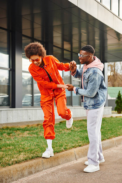 Cheerful and trendy young african american man in sunglasses holding hands of best friend walking on border near building on urban street, stylish friends enjoying company concept