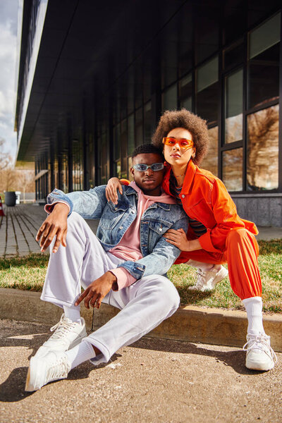 Full length of trendy and confident african american best friends in sunglasses looking at camera while sitting on border on blurred urban street at background, stylish friends enjoying company