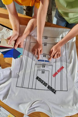 Top view of young african american designer holding printing layer near t-shirt and colleague working with color swatches on table in print studio, thriving small enterprise concept clipart