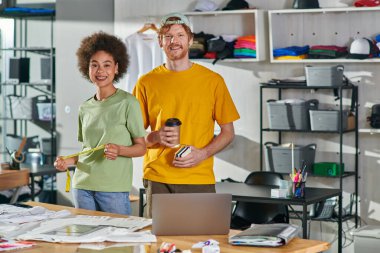 Positive multiethnic craftspeople holding coffee to go and tape measure while looking at camera and standing near laptop and clothes on table in print studio, start-up innovation concept clipart
