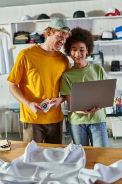 Smiling young african american designer using laptop near colleague with printing layer and clothes on table in blurred print studio at background, self-employment opportunity concept clipart