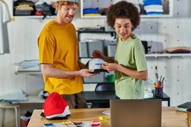 Baskı stüdyosundaki giysi örneklerinin yakınındaki dizüstü bilgisayara bakarken, baskı katmanı ve snapback ile çalışan gülümseyen çok ırklı genç tasarımcılar, sürdürülebilir iş modeli kavramı