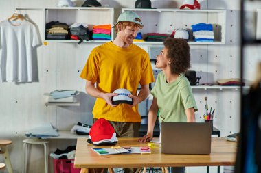Baskı stüdyosundaki kumaş örneklerinin yanında snapback ve laptop ile çalışırken konuşan pozitif genç tasarımcılar, sürdürülebilir iş modeli konsepti
