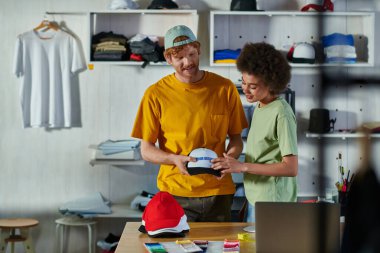 Gülümseyen çok ırklı genç tasarımcılar ellerinde baskı katmanı ve snapback tutarak baskı stüdyosunda dizüstü bilgisayar ve kumaş örnekleri üzerinde çalışırken, sürdürülebilir iş modeli konsepti