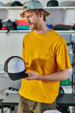 Genç kızıl saçlı zanaatkar projede çalışırken snapback yapıyor ve arka planda bulanık baskı stüdyosunda duruyor, küçük işletme esnekliği konsepti.