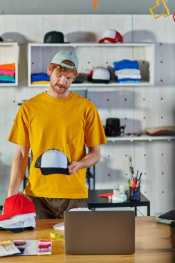 Genç kızıl saçlı zanaatkar dizüstü bilgisayar ve kumaş örnekleri üzerinde çalışırken snapback tutuyor ve bulanık baskı stüdyosunda duruyor, küçük işletme dayanıklılığı konsepti
