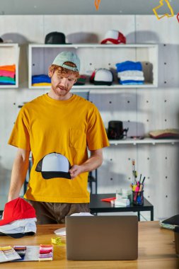 Young redhead craftsman holding snapback while working on project near laptop and cloth samples on table and standing in blurred print studio, small business resilience concept clipart