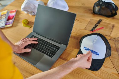 Tasarımcı görüşü boş ekranlı dizüstü bilgisayar kullanıyor ve baskı stüdyosundaki bulanık masanın yanında baskı katmanı tutuyor. 