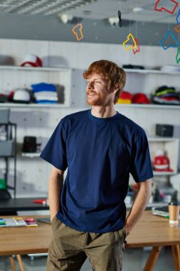 Genç kızıl saçlı tasarımcı ceplerinde el ele tutuşuyor ve bulanık baskı stüdyosunda çalışma masasının yanında duruyor. 