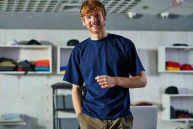 Portrait of positive young redhead craftsman in casual clothes looking at camera while posing and standing in blurred print studio, self-made success concept  clipart