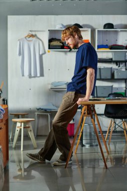 Side view of young redhead craftsman in casual clothes standing near smartphone wooden working table in print studio at background, self-made success concept  clipart