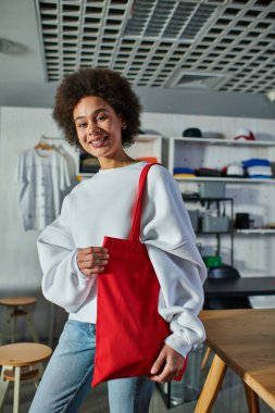 Cheerful young african american craftswoman holding shoulder bag and looking at camera while standing in blurred print studio, enthusiastic business owner working in workshop clipart