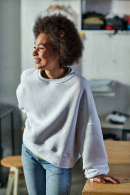 Tişört ve kot pantolon giymiş neşeli Afro-Amerikan zanaatkar kadın portresi. Bulanık baskı stüdyosunda çalışma masasının yanında duruyor.
