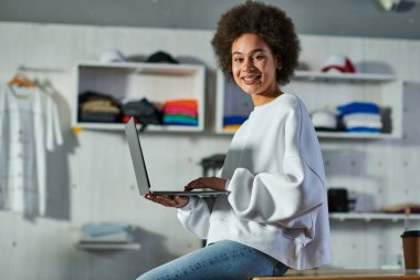 Sweatshirt 'lü ve kot pantolonlu neşeli Afro-Amerikalı kadın. Baskı stüdyosundaki masaya çıkmak için kahve yanındaki bilgisayarı kullanırken kameraya bakıyor.