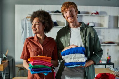 Smiling young multiethnic craftspeople holding clothes while standing and working together in blurred print studio at background, ambitious young entrepreneurs concept  clipart