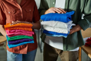 Cropped view of young interracial craftspeople holding clothes while standing next to each other and working in print studio, ambitious young entrepreneurs concept  clipart