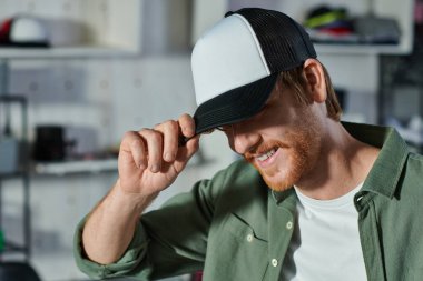 Arka planda bulanık baskı stüdyosunda çalışırken ve çalışırken snapback takan gülümseyen kızıl saçlı genç bir zanaatkar, müşteri odaklı girişimci konsepti 