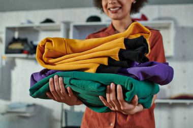 Cropped view of young blurred african american craftswoman holding clothes while standing and working in print studio at background, customer-focused entrepreneur concept  clipart